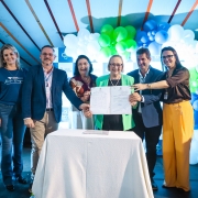 Foto posada na solenidade, com seis pessoas de p&eacute;, exibindo para a c&acirc;mera um documento assinado. Elas est&atilde;o atr&aacute;s de uma mesa e na frente de enfeites de bal&atilde;o. Todas sorriem.