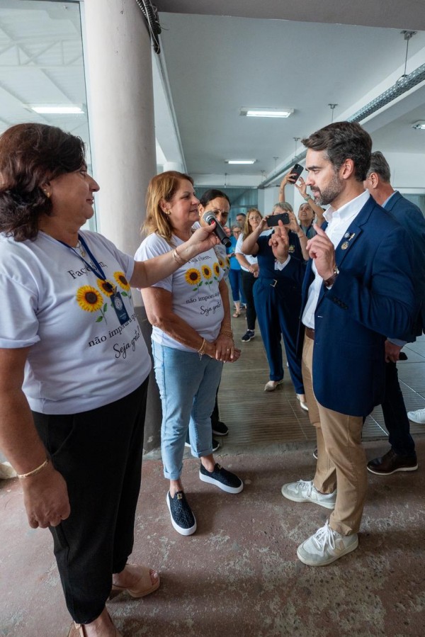 A fotografia captura um momento de diálogo entre o governador do Rio Grande do Sul, Eduardo Leite, e um grupo de mulheres em um ambiente interno amplo, possivelmente o pátio ou corredor de uma instituição pública.

Elementos Centrais
Eduardo Leite (Direita): O governador, um homem branco de cabelos e barba castanhos, está de perfil voltado para a esquerda. Ele veste um blazer azul-marinho, camisa branca sem gravata e calça de sarja bege. Ele gesticula com a mão direita levantada enquanto fala, demonstrando uma atitude explicativa ou enfática.

As Interlocutoras (Esquerda): Três mulheres estão em destaque à frente dele. Duas delas vestem camisetas brancas idênticas com uma estampa de três girassóis e a frase: "Faça o bem, não importa a quem. Seja grato!".

A mulher mais à esquerda segura um microfone próximo ao governador, indicando que a fala dele está sendo amplificada ou gravada.

A mulher ao centro ouve atentamente com as mãos cruzadas à frente do corpo.

Composição e Fundo
Público e Registro: Ao fundo, outras pessoas observam a cena. Algumas mulheres aparecem segurando celulares para fotografar ou filmar o encontro, reforçando o caráter oficial e público do evento.

Ambiente: O local possui colunas cilíndricas brancas e um teto alto com luminárias fluorescentes. O chão é de cimento com tons avermelhados e marcas de uso. A profundidade de campo mostra um corredor que se estende ao fundo, conferindo uma sensação de amplitude.

Perspectiva: A foto é tirada de um ângulo ligeiramente baixo, o que dá destaque às figuras centrais da interação.

Atmosfera
A cena transmite uma sensação de proximidade e diálogo direto. O contato visual entre o governador e as mulheres sugere uma conversa séria, porém acessível, típica de vistorias ou encontros comunitários.