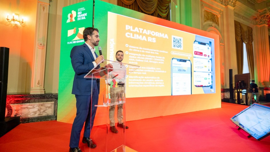 O governador e o vice-governador est&atilde;o posicionados ao lado de um p&uacute;lpito transparente, de frente para uma tela grande que exibe a apresenta&ccedil;&atilde;o &ldquo;Plataforma Clima RS&rdquo;, acompanhada de um QR Code e da interface do sistema mostrada em um dispositivo m&oacute;vel. 