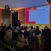 Foto mostra palestrante no palco mostrando dados numa tela, enquanto plateia acompanha apresenta&ccedil;&atilde;o.