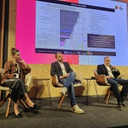 Painel de discuss&atilde;o em evento institucional, com tr&ecirc;s participantes sentados em poltronas no palco. Ao fundo, um tel&atilde;o exibe gr&aacute;ficos e informa&ccedil;&otilde;es sobre startups. 