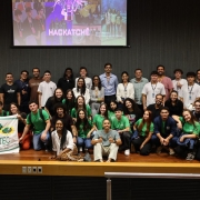 Esta &eacute; uma descri&ccedil;&atilde;o detalhada da imagem para fins de acessibilidade:

A fotografia mostra um grande grupo de jovens e alguns adultos reunidos no palco de um audit&oacute;rio para o an&uacute;ncio das equipes finalistas do HackaTch&ecirc; 2026.

Elementos Centrais e Composi&ccedil;&atilde;o
O Grupo: Cerca de 30 pessoas posam para a foto. O grupo &eacute; diversificado e demonstra entusiasmo. Muitos jovens vestem camisetas verdes com o logotipo do evento, enquanto outros usam roupas casuais. Eles est&atilde;o organizados em diferentes n&iacute;veis: alguns em p&eacute; ao fundo, outros sentados ou agachados &agrave; frente.

Destaque Visual (Esquerda): Dois jovens seguram uma bandeira branca da ETEC (Escola T&eacute;cnica Estadual Celeiro), de Bom Progresso - RS. A bandeira cont&eacute;m ilustra&ccedil;&otilde;es coloridas que remetem &agrave; agricultura e ao ensino t&eacute;cnico.

Proje&ccedil;&atilde;o ao Fundo: No alto, uma tela de proje&ccedil;&atilde;o exibe a identidade visual do evento com o nome "HACKATCH&Ecirc;" em letras garrafais brancas, sob a imagem de uma l&acirc;mpada estilizada (s&iacute;mbolo de ideia/inova&ccedil;&atilde;o) feita de circuitos de luz roxa.

Ambiente do Audit&oacute;rio
Palco: O piso &eacute; de madeira clara (tablado), e a parte frontal do palco tem um acabamento em madeira com detalhes met&aacute;licos.

Paredes: O fundo do palco &eacute; composto por pain&eacute;is ac&uacute;sticos ripados em tom cinza-escuro, conferindo um aspecto moderno e profissional ao ambiente.

Ilumina&ccedil;&atilde;o: A luz &eacute; focada no grupo, destacando as express&otilde;es de alegria e conquista dos participantes.

Atmosfera
A imagem transmite um clima de celebra&ccedil;&atilde;o, inova&ccedil;&atilde;o e protagonismo juvenil. &Eacute; um registro de encerramento de uma etapa importante, onde o esp&iacute;rito de equipe e o orgulho pela representa&ccedil;&atilde;o de suas escolas s&atilde;o evidentes.