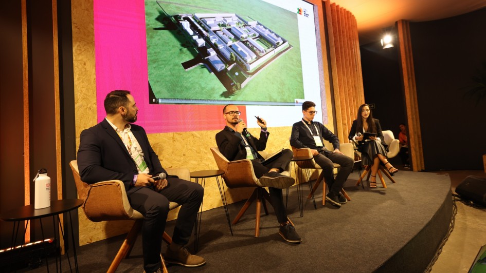 Foto mostra palestrante sentado, olhando para tela onde h&aacute; uma foto de um projeto de uma unidade prisional. Outros tr&ecirc;s palestrantes acompanham a fala, tamb&eacute;m sentados. 