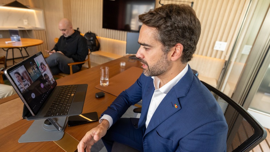 Homem de terno azul participa de uma videoconfer&ecirc;ncia em um tablet apoiado sobre a mesa. Ele gesticula enquanto fala. Ao fundo, outro homem sentado mexe no celular, em uma sala moderna com paredes de madeira e uma TV.