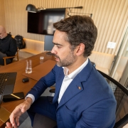 Homem de terno azul participa de uma videoconfer&ecirc;ncia em um tablet apoiado sobre a mesa. Ele gesticula enquanto fala. Ao fundo, outro homem sentado mexe no celular, em uma sala moderna com paredes de madeira e uma TV.