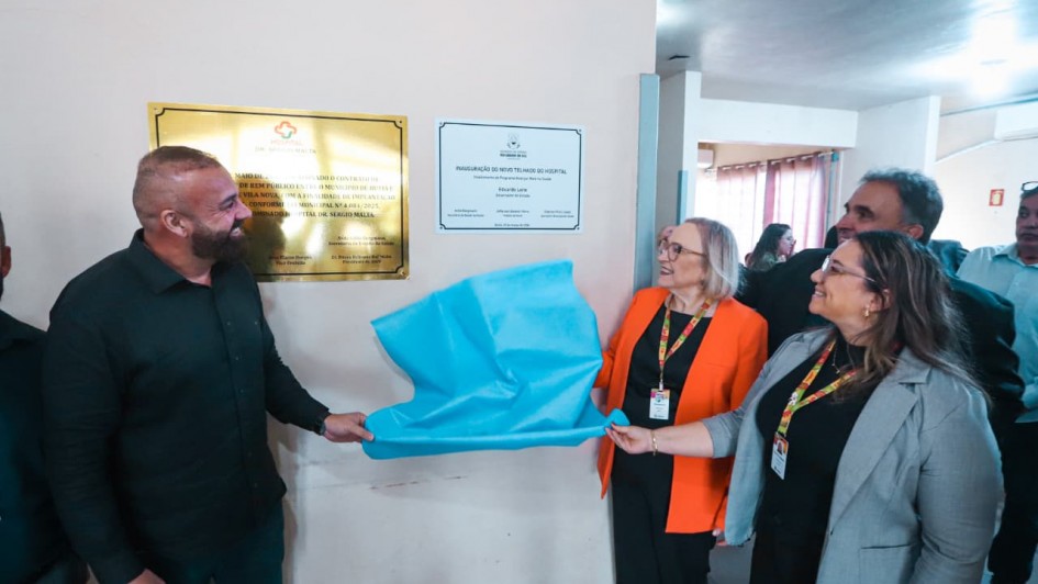 A fotografia registra o momento solene e festivo do descerramento de duas placas de inaugura&ccedil;&atilde;o em uma unidade de sa&uacute;de, com a presen&ccedil;a de autoridades estaduais e municipais.

A A&ccedil;&atilde;o Central (Descerramento)
Pessoas: Quatro pessoas est&atilde;o em destaque &agrave; frente de uma parede branca.

Esquerda: Um homem de barba, vestindo uma camisa preta e cal&ccedil;a escura, sorri e segura uma das pontas de um tecido azul-turquesa que cobria as placas.

Direita: Duas mulheres e um homem est&atilde;o em p&eacute; ao lado dele, tamb&eacute;m sorrindo e participando do descerramento.

A mulher ao centro veste um blazer laranja vibrante sobre uma blusa preta, com um crach&aacute; colorido no pesco&ccedil;o.

A mulher mais &agrave; direita veste um blazer cinza-claro e segura a outra ponta do tecido azul.

O Tecido: Um pano azul-turquesa claro, que cobria as placas, est&aacute; sendo puxado e dobra-se no ar, revelando as placas.

As Placas (Fundo)
Placa da Esquerda: Uma placa dourada retangular e reluzente. O texto &eacute; mais denso e formal, incluindo logotipos do HOSPITAL BOM PASTOR DE GRAVATA&Iacute; e men&ccedil;&otilde;es a atos municipais e estaduais de coopera&ccedil;&atilde;o.

Placa da Direita: Uma placa branca emoldurada de forma mais simples. &Eacute; a placa principal de inaugura&ccedil;&atilde;o, com o bras&atilde;o do Estado do Rio Grande do Sul no topo. O texto principal &eacute;: "INAUGURA&Ccedil;&Atilde;O DO NOVO TELHADO DO HOSPITAL", especificando que o investimento &eacute; do Programa Avan&ccedil;ar na Sa&uacute;de do Governo do Estado. Abaixo, l&ecirc;-se o nome do governador "Eduardo Leite" e de outros secret&aacute;rios estaduais.

Ambiente e Outras Pessoas
Ambiente: A parede &eacute; branca com um barrado de m&aacute;rmore esverdeado na base. Ao fundo, v&ecirc;-se um corredor amplo com piso de cimento, colunas brancas e janelas com cortinas, al&eacute;m de mais pessoas que observam a cerim&ocirc;nia. A ilumina&ccedil;&atilde;o &eacute; de teto, conferindo um tom claro e natural &agrave; cena.

P&uacute;blico: &Agrave; direita, atr&aacute;s da mulher de cinza, um homem de terno preto e outra pessoa assistem &agrave; cena. Mais ao fundo, distinguem-se outras silhuetas e um extintor de inc&ecirc;ndio na parede.

Atmosfera
A cena transmite alegria, orgulho e coopera&ccedil;&atilde;o entre as diferentes esferas de governo para a melhoria de uma infraestrutura p&uacute;blica de sa&uacute;de. &Eacute; um registro de uma conquista entregue &agrave; comunidade.