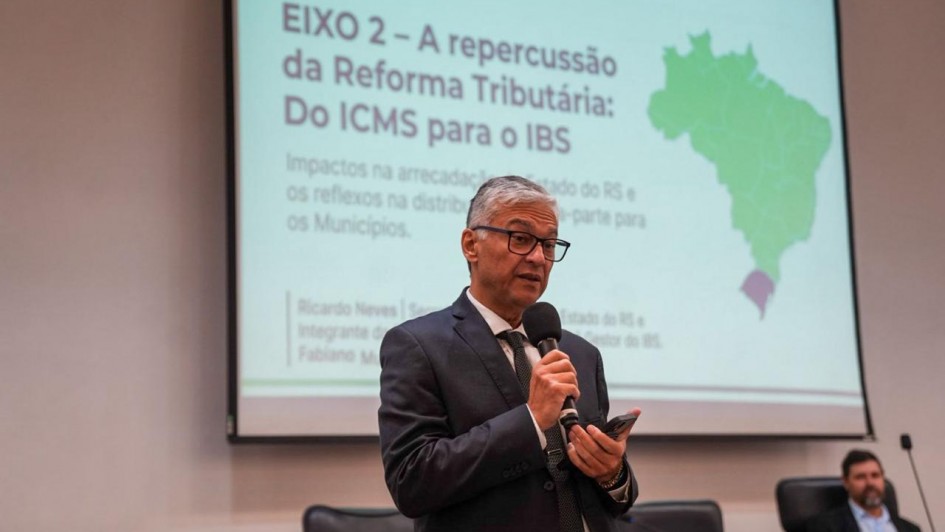 Foto mostra palestrante falando em p&eacute;, com microfone na m&atilde;o. Atr&aacute;s dele, aparece uma tela com uma apresenta&ccedil;&atilde;o e o mapa do Brasil.