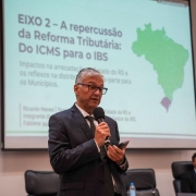 Foto mostra palestrante falando em p&eacute;, com microfone na m&atilde;o. Atr&aacute;s dele, aparece uma tela com uma apresenta&ccedil;&atilde;o e o mapa do Brasil.