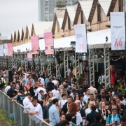 A fotografia a&eacute;rea, capturada em um &acirc;ngulo levemente elevado, mostra a intensa movimenta&ccedil;&atilde;o de p&uacute;blico no Cais Embarcadero, em Porto Alegre, durante o South Summit Brazil 2026.

O P&uacute;blico e a Movimenta&ccedil;&atilde;o
Multid&atilde;o: Centenas de pessoas de perfis diversos circulam por um amplo corredor a c&eacute;u aberto. A maioria veste roupas casuais ou profissionais (estilo business casual) e muitos portam crach&aacute;s oficiais do evento pendurados no pesco&ccedil;o. O clima &eacute; de grande efervesc&ecirc;ncia e circula&ccedil;&atilde;o constante.

Corredor Principal: O fluxo de pessoas ocorre entre uma grade de prote&ccedil;&atilde;o &agrave; esquerda (que separa a &aacute;rea do Gua&iacute;ba) e uma estrutura de tendas brancas e estandes &agrave; direita.

Estruturas e Sinaliza&ccedil;&atilde;o
Banners e Totens: Diversas bandeiras verticais e totens de sinaliza&ccedil;&atilde;o em tons de branco, rosa e azul est&atilde;o espalhados pelo trajeto, orientando o p&uacute;blico para &aacute;reas como:

Food Court 2 (Pra&ccedil;a de alimenta&ccedil;&atilde;o)

Music Stage (Palco de m&uacute;sica)

Toilet (Banheiros)

Experience VIP Lounge by PUCRS

Patroc&iacute;nios e Parcerias: Um grande totem &agrave; esquerda exibe os logotipos dos principais realizadores e parceiros, com destaque para o Governo do Estado do Rio Grande do Sul, SEBRAE, FIERGS e a prefeitura de Porto Alegre.

Arquitetura do Cais: Ao fundo, alinham-se os hist&oacute;ricos telhados em formato de "serra" (shed) dos armaz&eacute;ns do Cais, que contrastam com as estruturas met&aacute;licas modernas e as tendas tempor&aacute;rias montadas para o evento tecnol&oacute;gico.

Ambiente e Composi&ccedil;&atilde;o
C&eacute;u e Ilumina&ccedil;&atilde;o: O dia parece nublado, com uma luz difusa que ilumina uniformemente a multid&atilde;o sem criar sombras fortes.

Perspectiva: A profundidade de campo mostra que o evento se estende por uma longa dist&acirc;ncia ao longo da orla, enfatizando a magnitude da infraestrutura montada.

Atmosfera
A imagem transmite uma sensa&ccedil;&atilde;o de vitalidade, conex&atilde;o e escala, posicionando o Rio Grande do Sul como um polo vibrante de inova&ccedil;&atilde;o, tecnologia e grandes encontros globais.