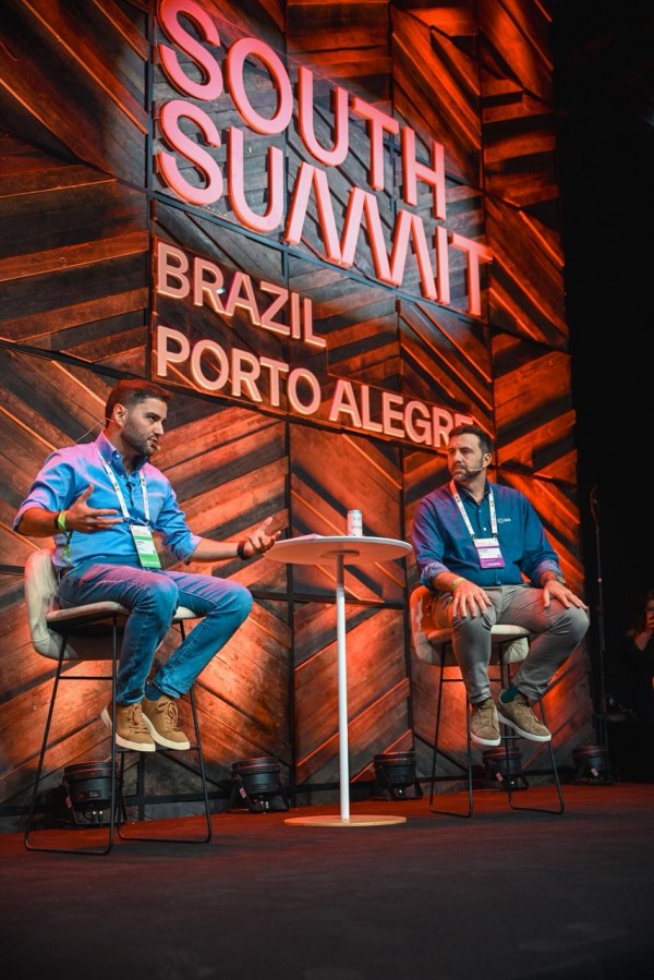 Cena de debate no South Summit Brazil, com dois participantes sentados no palco e uma mesa alta entre eles. O fundo destaca o nome do evento em letras iluminadas, com iluminação cênica e ambiente corporativo.