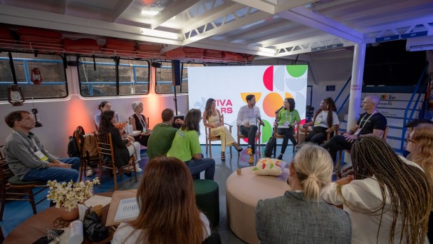 Esta &eacute; uma descri&ccedil;&atilde;o detalhada da imagem para fins de acessibilidade:

A fotografia registra uma roda de conversa sobre iniciativas de impacto social apoiadas pelo BRDE, realizada em um ambiente singular: o interior de uma embarca&ccedil;&atilde;o atracada no Cais Embarcadero, durante o South Summit Brazil 2026.

O Painel e a Din&acirc;mica
Disposi&ccedil;&atilde;o: Cerca de dez pessoas est&atilde;o organizadas em um c&iacute;rculo, promovendo um ambiente de debate pr&oacute;ximo e horizontal.

Participantes em Destaque: Ao centro-fundo, quatro debatedores est&atilde;o sentados em cadeiras de madeira. Uma mulher com camiseta verde lim&atilde;o fala ao microfone, enquanto os demais &mdash; um homem de camisa branca e cal&ccedil;a escura e duas mulheres (uma de amarelo e outra de camiseta branca) &mdash; ouvem atentamente.

Plateia Pr&oacute;xima: Outras pessoas ocupam cadeiras laterais e bancos acolhedores (pufes) em primeiro plano, participando da conversa em um formato de "aqu&aacute;rio" ou roda aberta.

O Ambiente (Interior do Barco)
Estrutura: O teto &eacute; baixo, com vigas met&aacute;licas brancas expostas, t&iacute;picas de estruturas navais. Nas laterais, veem-se janelas retangulares com molduras pretas que mostram a parede de pedra do cais do lado de fora.

Decora&ccedil;&atilde;o Tem&aacute;tica: Lumin&aacute;rias que lembram lampi&otilde;es antigos est&atilde;o penduradas ou fixadas nas paredes, refor&ccedil;ando a est&eacute;tica n&aacute;utica. Uma tela de LED ao fundo exibe a marca "VIVA RS" envolta em grafismos coloridos (c&iacute;rculos e semic&iacute;rculos em vermelho, verde e amarelo).

Mobili&aacute;rio: Al&eacute;m das cadeiras de madeira, h&aacute; mesinhas de apoio com flores, garrafas de &aacute;gua e materiais informativos. Pufes redondos em tons past&eacute;is e verde escuro oferecem assentos informais.

Ilumina&ccedil;&atilde;o e Composi&ccedil;&atilde;o
Luz: A ilumina&ccedil;&atilde;o &eacute; suave e focada nos participantes, com pontos de luz quente (alaranjada) nas paredes, contrastando com a claridade branca vinda do teto e da tela de LED.

Perspectiva: A foto &eacute; tirada de um &acirc;ngulo aberto, capturando tanto as cabe&ccedil;as das pessoas em primeiro plano (vistas de costas) quanto o painel principal, o que coloca o observador "dentro" da conversa.

Atmosfera
A cena transmite uma sensa&ccedil;&atilde;o de intimidade, colabora&ccedil;&atilde;o e inova&ccedil;&atilde;o social. O uso de um barco como palco para debater o futuro e o impacto social reflete o esp&iacute;rito criativo do evento em Porto Alegre.