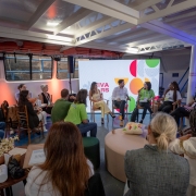 Esta &eacute; uma descri&ccedil;&atilde;o detalhada da imagem para fins de acessibilidade:

A fotografia registra uma roda de conversa sobre iniciativas de impacto social apoiadas pelo BRDE, realizada em um ambiente singular: o interior de uma embarca&ccedil;&atilde;o atracada no Cais Embarcadero, durante o South Summit Brazil 2026.

O Painel e a Din&acirc;mica
Disposi&ccedil;&atilde;o: Cerca de dez pessoas est&atilde;o organizadas em um c&iacute;rculo, promovendo um ambiente de debate pr&oacute;ximo e horizontal.

Participantes em Destaque: Ao centro-fundo, quatro debatedores est&atilde;o sentados em cadeiras de madeira. Uma mulher com camiseta verde lim&atilde;o fala ao microfone, enquanto os demais &mdash; um homem de camisa branca e cal&ccedil;a escura e duas mulheres (uma de amarelo e outra de camiseta branca) &mdash; ouvem atentamente.

Plateia Pr&oacute;xima: Outras pessoas ocupam cadeiras laterais e bancos acolhedores (pufes) em primeiro plano, participando da conversa em um formato de "aqu&aacute;rio" ou roda aberta.

O Ambiente (Interior do Barco)
Estrutura: O teto &eacute; baixo, com vigas met&aacute;licas brancas expostas, t&iacute;picas de estruturas navais. Nas laterais, veem-se janelas retangulares com molduras pretas que mostram a parede de pedra do cais do lado de fora.

Decora&ccedil;&atilde;o Tem&aacute;tica: Lumin&aacute;rias que lembram lampi&otilde;es antigos est&atilde;o penduradas ou fixadas nas paredes, refor&ccedil;ando a est&eacute;tica n&aacute;utica. Uma tela de LED ao fundo exibe a marca "VIVA RS" envolta em grafismos coloridos (c&iacute;rculos e semic&iacute;rculos em vermelho, verde e amarelo).

Mobili&aacute;rio: Al&eacute;m das cadeiras de madeira, h&aacute; mesinhas de apoio com flores, garrafas de &aacute;gua e materiais informativos. Pufes redondos em tons past&eacute;is e verde escuro oferecem assentos informais.

Ilumina&ccedil;&atilde;o e Composi&ccedil;&atilde;o
Luz: A ilumina&ccedil;&atilde;o &eacute; suave e focada nos participantes, com pontos de luz quente (alaranjada) nas paredes, contrastando com a claridade branca vinda do teto e da tela de LED.

Perspectiva: A foto &eacute; tirada de um &acirc;ngulo aberto, capturando tanto as cabe&ccedil;as das pessoas em primeiro plano (vistas de costas) quanto o painel principal, o que coloca o observador "dentro" da conversa.

Atmosfera
A cena transmite uma sensa&ccedil;&atilde;o de intimidade, colabora&ccedil;&atilde;o e inova&ccedil;&atilde;o social. O uso de um barco como palco para debater o futuro e o impacto social reflete o esp&iacute;rito criativo do evento em Porto Alegre.