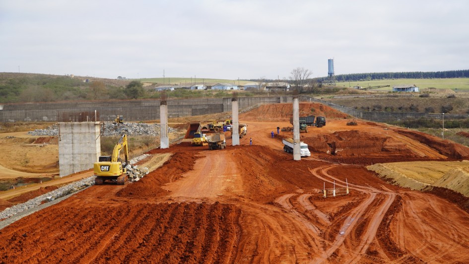 Imagem de uma grande &aacute;rea em obras, com maquin&aacute;rio como retroescavadeiras e ca&ccedil;ambas circulando na &aacute;rea. No centro, tr&ecirc;s grandes pilares de concreto