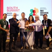 Um grupo de seis pessoas se posiciona ao redor de uma pequena mesa segurando um documento. Ao fundo, um painel com as logo marcas do South Summit Brazil e do governo do RS.