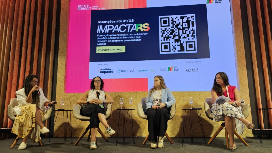 Quatro mulheres sentadas em cadeiras dispostas em um palco. Aoo fundo, um banner com a logomarca da ImpactaRS