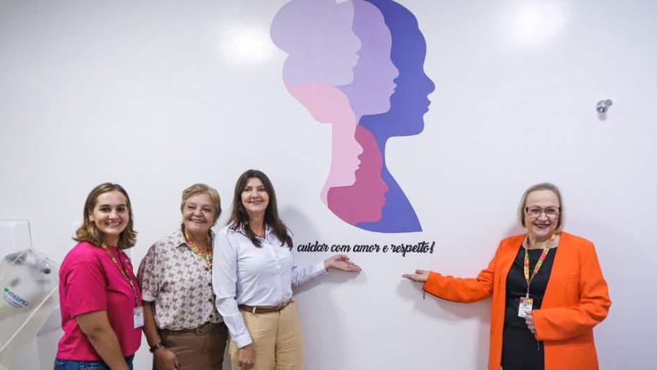 A fotografia registra a inaugura&ccedil;&atilde;o do espa&ccedil;o SERMulher RS em Ven&acirc;ncio Aires, destacando quatro mulheres sorridentes em frente a um painel tem&aacute;tico em uma parede branca.

Elementos Centrais e Pessoas
O Grupo: Quatro mulheres est&atilde;o posicionadas lado a lado, em uma composi&ccedil;&atilde;o que transmite uni&atilde;o e celebra&ccedil;&atilde;o.

&Agrave; esquerda: Uma jovem mulher de camiseta rosa vibrante e cal&ccedil;a jeans.

Segunda da esquerda: Uma mulher madura com uma camisa estampada em tons de bege e marrom.

Terceira da esquerda: Uma mulher de camisa branca e cal&ccedil;a bege, que aponta com a m&atilde;o direita para o logotipo na parede.

&Agrave; direita: Uma mulher de &oacute;culos, vestindo um blazer laranja intenso sobre blusa preta, tamb&eacute;m aponta para o logotipo. Ela utiliza um crach&aacute; oficial com cord&atilde;o colorido.

O Painel (Parede): No centro da parede, h&aacute; uma arte gr&aacute;fica composta por silhuetas de perfis femininos sobrepostos em diferentes tons de lil&aacute;s, rosa e roxo. Acima da arte, l&ecirc;-se o texto: "SER MULHER..." e, abaixo, a frase em fonte cursiva: "cuidar com amor e respeito!".

Ambiente e Composi&ccedil;&atilde;o
Cen&aacute;rio: O ambiente &eacute; interno, com paredes brancas e ilumina&ccedil;&atilde;o clara e direta, t&iacute;pica de uma unidade de sa&uacute;de ou centro de atendimento. &Agrave; extrema esquerda, nota-se parte de um equipamento m&eacute;dico ou odontol&oacute;gico coberto por um pl&aacute;stico protetor.

Enquadramento: A foto &eacute; frontal, centralizando as autoridades e o s&iacute;mbolo do novo servi&ccedil;o, focando na identidade visual do programa governamental.

Atmosfera
A imagem transmite um sentimento de acolhimento, realiza&ccedil;&atilde;o e compromisso com as pol&iacute;ticas p&uacute;blicas voltadas &agrave;s mulheres. As cores vibrantes das roupas (rosa e laranja) contrastam com o fundo branco, refor&ccedil;ando o tom positivo do evento.