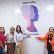 A fotografia registra a inaugura&ccedil;&atilde;o do espa&ccedil;o SERMulher RS em Ven&acirc;ncio Aires, destacando quatro mulheres sorridentes em frente a um painel tem&aacute;tico em uma parede branca.

Elementos Centrais e Pessoas
O Grupo: Quatro mulheres est&atilde;o posicionadas lado a lado, em uma composi&ccedil;&atilde;o que transmite uni&atilde;o e celebra&ccedil;&atilde;o.

&Agrave; esquerda: Uma jovem mulher de camiseta rosa vibrante e cal&ccedil;a jeans.

Segunda da esquerda: Uma mulher madura com uma camisa estampada em tons de bege e marrom.

Terceira da esquerda: Uma mulher de camisa branca e cal&ccedil;a bege, que aponta com a m&atilde;o direita para o logotipo na parede.

&Agrave; direita: Uma mulher de &oacute;culos, vestindo um blazer laranja intenso sobre blusa preta, tamb&eacute;m aponta para o logotipo. Ela utiliza um crach&aacute; oficial com cord&atilde;o colorido.

O Painel (Parede): No centro da parede, h&aacute; uma arte gr&aacute;fica composta por silhuetas de perfis femininos sobrepostos em diferentes tons de lil&aacute;s, rosa e roxo. Acima da arte, l&ecirc;-se o texto: "SER MULHER..." e, abaixo, a frase em fonte cursiva: "cuidar com amor e respeito!".

Ambiente e Composi&ccedil;&atilde;o
Cen&aacute;rio: O ambiente &eacute; interno, com paredes brancas e ilumina&ccedil;&atilde;o clara e direta, t&iacute;pica de uma unidade de sa&uacute;de ou centro de atendimento. &Agrave; extrema esquerda, nota-se parte de um equipamento m&eacute;dico ou odontol&oacute;gico coberto por um pl&aacute;stico protetor.

Enquadramento: A foto &eacute; frontal, centralizando as autoridades e o s&iacute;mbolo do novo servi&ccedil;o, focando na identidade visual do programa governamental.

Atmosfera
A imagem transmite um sentimento de acolhimento, realiza&ccedil;&atilde;o e compromisso com as pol&iacute;ticas p&uacute;blicas voltadas &agrave;s mulheres. As cores vibrantes das roupas (rosa e laranja) contrastam com o fundo branco, refor&ccedil;ando o tom positivo do evento.