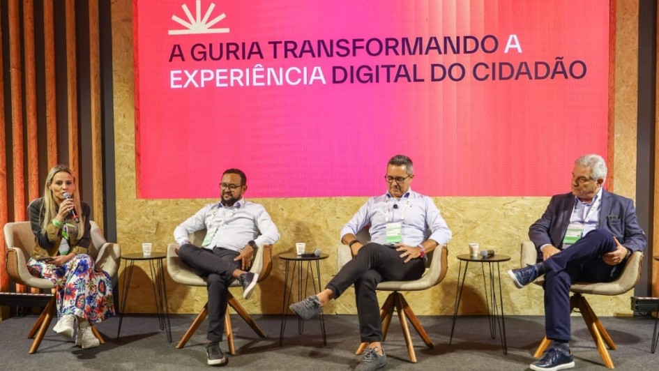 A foto registra um painel no South Summit Brazil, com quatro participantes sentados em poltronas diante de um grande tel&atilde;o rosa que exibe o texto: &ldquo;A guria transformando a experi&ecirc;ncia digital do cidad&atilde;o&rdquo;. O palco tem decora&ccedil;&atilde;o em madeira vertical e ilumina&ccedil;&atilde;o direcionada, enquanto a plateia assiste ao debate nas primeiras fileiras.