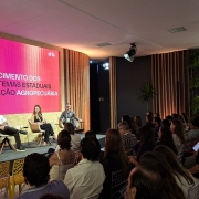 Esta &eacute; uma descri&ccedil;&atilde;o detalhada da imagem para fins de acessibilidade:

A fotografia registra um painel de discuss&atilde;o durante o South Summit Brazil 2026, focado no setor prim&aacute;rio e inova&ccedil;&atilde;o.

O Palco e a Apresenta&ccedil;&atilde;o
Painelistas: Quatro pessoas (tr&ecirc;s homens e uma mulher) est&atilde;o sentadas em poltronas girat&oacute;rias de cor clara sobre um palco baixo. Eles est&atilde;o dispostos em semic&iacute;rculo, voltados uns para os outros e para a plateia, em um clima de conversa informal.

O Tel&atilde;o: Um grande painel de LED ao fundo exibe um slide com fundo rosa vibrante e letras brancas, onde se l&ecirc;: "FORTALECIMENTO DOS ECOSSISTEMAS ESTADUAIS DE INOVA&Ccedil;&Atilde;O AGROPECU&Aacute;RIA". No canto superior esquerdo do tel&atilde;o, aparece o logotipo do South Summit Brazil Porto Alegre.

Cenografia: O palco &eacute; emoldurado por pain&eacute;is de madeira clara e ripas verticais que criam um efeito de luz e sombra.

O Ambiente e o P&uacute;blico
Audit&oacute;rio: A plateia est&aacute; lotada, com dezenas de pessoas sentadas em cadeiras transparentes e coloridas (amarelas e vermelhas). O p&uacute;blico est&aacute; de costas para a c&acirc;mera, com o olhar atento ao palco.

Decora&ccedil;&atilde;o Lateral: &Agrave; direita da imagem, destaca-se uma parede decorada com samambaias verdes dispostas em uma grade met&aacute;lica, iluminada por tubos de LED branco em formatos geom&eacute;tricos, similar &agrave; cenografia de outros pain&eacute;is do evento.

Ilumina&ccedil;&atilde;o: O ambiente possui uma ilumina&ccedil;&atilde;o c&ecirc;nica sofisticada, com refletores de luz negra e branca pendurados no teto, criando um contraste moderno entre o palco iluminado e a plateia na penumbra.

Atmosfera
A cena transmite uma sensa&ccedil;&atilde;o de conhecimento compartilhado e foco tecnol&oacute;gico. A imagem refor&ccedil;a a import&acirc;ncia do agroneg&oacute;cio dentro do ecossistema de inova&ccedil;&atilde;o global, unindo especialistas para debater o futuro do setor no Rio Grande do Sul.