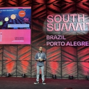Homem em um grande palco de p&eacute;, em palestra. Atr&aacute;s dele, uma parede decorada, com luzes e a logomarca South Summit Brazil