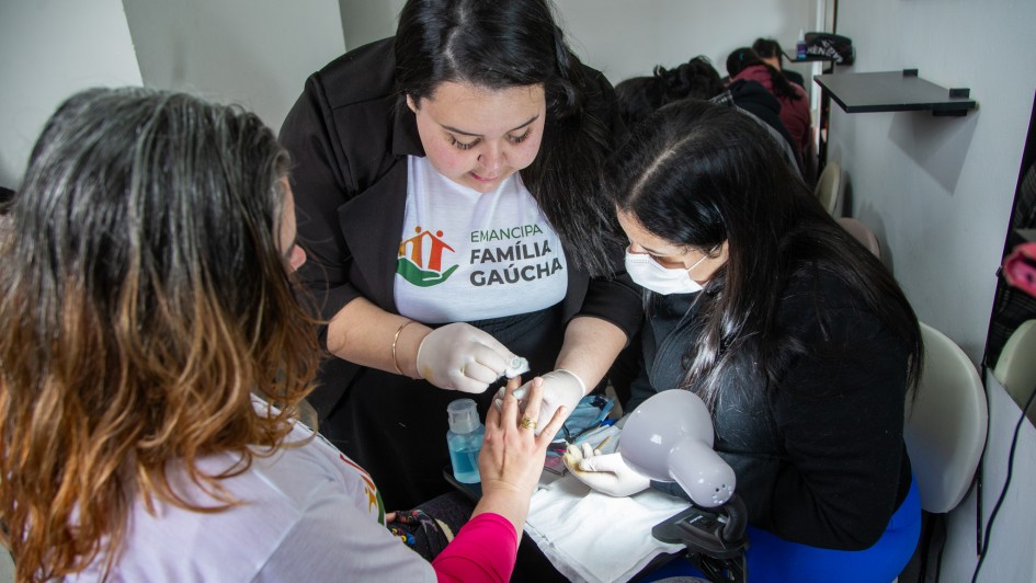 Foto mostra uma aluna de manicure fazendo as unhas de uma mulher, enquanto instrutora com a camiseta do projeto Emancipa Fam&iacute;lia orienta como fazer.