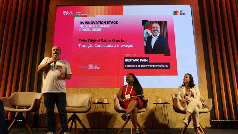Esta &eacute; uma descri&ccedil;&atilde;o detalhada da imagem para fins de acessibilidade:

A fotografia registra um painel de discuss&atilde;o no palco do RS Innovation Stage, durante o South Summit Brazil 2026, com foco no desenvolvimento rural e inova&ccedil;&atilde;o digital.

O Palco e a Apresenta&ccedil;&atilde;o
Participantes: Tr&ecirc;s pessoas est&atilde;o no palco.

&Agrave; esquerda, um homem de camiseta branca e cal&ccedil;a jeans est&aacute; em p&eacute;, falando ao microfone e gesticulando.

Ao centro e &agrave; direita, duas mulheres est&atilde;o sentadas em poltronas girat&oacute;rias cinzas, ouvindo atentamente. A mulher ao centro veste um vestido vermelho vibrante e a da direita veste tons de bege e branco.

O Tel&atilde;o: O grande painel de LED ao fundo possui um design em tons de rosa e vermelho. Ele exibe o t&iacute;tulo: "Feira Digital Sabor Ga&uacute;cho: Tradi&ccedil;&atilde;o Conectada &agrave; inova&ccedil;&atilde;o".

Destaque no Tel&atilde;o: H&aacute; uma foto e o nome do palestrante Gustavo Paim, identificado como Secret&aacute;rio de Desenvolvimento Rural. O logotipo do Governo do Estado do Rio Grande do Sul aparece no canto superior direito.

Cenografia: O palco &eacute; decorado com pain&eacute;is de madeira clara (OSB) na base e ripas de madeira verticais nas laterais, seguindo o padr&atilde;o visual das arenas de inova&ccedil;&atilde;o do evento.

Ambiente e Ilumina&ccedil;&atilde;o
A ilumina&ccedil;&atilde;o &eacute; focada nos palestrantes, com refletores de luz quente vindos do teto e das laterais.

O ch&atilde;o do palco &eacute; revestido por um carpete cinza escuro.

Entre as poltronas, h&aacute; pequenas mesas de apoio pretas com copos de &aacute;gua.

Atmosfera
A cena transmite um clima de entusiasmo e moderniza&ccedil;&atilde;o. O painel simboliza o esfor&ccedil;o do governo em digitalizar e promover os produtos da agricultura familiar e regional (Sabor Ga&uacute;cho) dentro de um ambiente de alta tecnologia e empreendedorismo global.