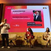Esta &eacute; uma descri&ccedil;&atilde;o detalhada da imagem para fins de acessibilidade:

A fotografia registra um painel de discuss&atilde;o no palco do RS Innovation Stage, durante o South Summit Brazil 2026, com foco no desenvolvimento rural e inova&ccedil;&atilde;o digital.

O Palco e a Apresenta&ccedil;&atilde;o
Participantes: Tr&ecirc;s pessoas est&atilde;o no palco.

&Agrave; esquerda, um homem de camiseta branca e cal&ccedil;a jeans est&aacute; em p&eacute;, falando ao microfone e gesticulando.

Ao centro e &agrave; direita, duas mulheres est&atilde;o sentadas em poltronas girat&oacute;rias cinzas, ouvindo atentamente. A mulher ao centro veste um vestido vermelho vibrante e a da direita veste tons de bege e branco.

O Tel&atilde;o: O grande painel de LED ao fundo possui um design em tons de rosa e vermelho. Ele exibe o t&iacute;tulo: "Feira Digital Sabor Ga&uacute;cho: Tradi&ccedil;&atilde;o Conectada &agrave; inova&ccedil;&atilde;o".

Destaque no Tel&atilde;o: H&aacute; uma foto e o nome do palestrante Gustavo Paim, identificado como Secret&aacute;rio de Desenvolvimento Rural. O logotipo do Governo do Estado do Rio Grande do Sul aparece no canto superior direito.

Cenografia: O palco &eacute; decorado com pain&eacute;is de madeira clara (OSB) na base e ripas de madeira verticais nas laterais, seguindo o padr&atilde;o visual das arenas de inova&ccedil;&atilde;o do evento.

Ambiente e Ilumina&ccedil;&atilde;o
A ilumina&ccedil;&atilde;o &eacute; focada nos palestrantes, com refletores de luz quente vindos do teto e das laterais.

O ch&atilde;o do palco &eacute; revestido por um carpete cinza escuro.

Entre as poltronas, h&aacute; pequenas mesas de apoio pretas com copos de &aacute;gua.

Atmosfera
A cena transmite um clima de entusiasmo e moderniza&ccedil;&atilde;o. O painel simboliza o esfor&ccedil;o do governo em digitalizar e promover os produtos da agricultura familiar e regional (Sabor Ga&uacute;cho) dentro de um ambiente de alta tecnologia e empreendedorismo global.