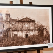 A imagem mostra outra fotografia histórica das ruínas missioneiras, em tom sépia, exibida em moldura preta sobre um cavalete. A antiga igreja de pedra aparece em estado de ruína, com paredes altas e detalhes arquitetônicos que evidenciam a grandiosidade do conjunto original e seu valor como patrimônio cultural.


