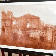 A imagem mostra uma fotografia em tom sépia das ruínas das Missões Jesuíticas Guaranis emoldurada e exposta sobre um cavalete. A construção de pedra aparece parcialmente tomada pela vegetação, com paredes desgastadas e aberturas irregulares, evidenciando o passar do tempo e reforçando o caráter histórico e patrimonial do conjunto arquitetônico retratado.

