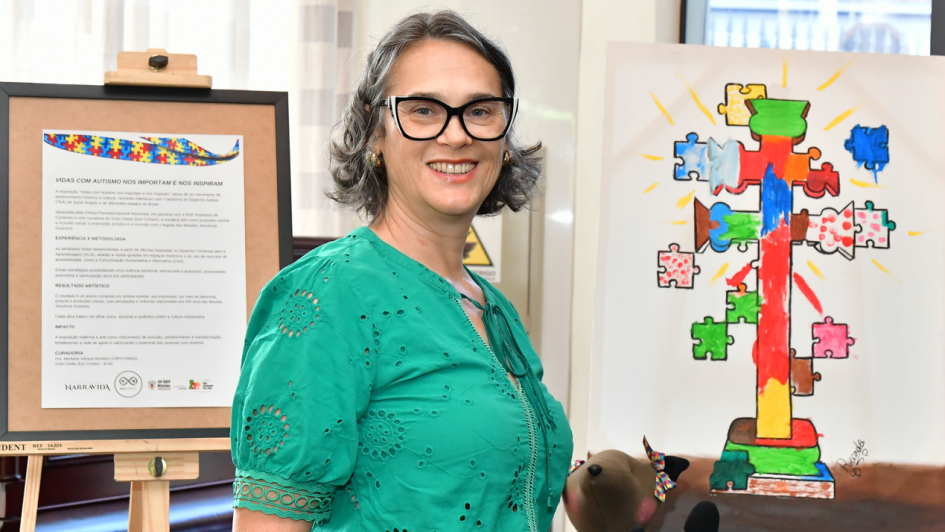 Na imagem, uma pessoa posa ao lado de obras da exposi&ccedil;&atilde;o &ldquo;Vidas com autismo nos importam e nos inspiram&rdquo;. Ao fundo, v&ecirc;-se um quadro com texto explicativo em suporte de madeira e, ao lado, uma pintura colorida que representa uma cruz formada por pe&ccedil;as de quebra-cabe&ccedil;a, associando simbolicamente inclus&atilde;o, diversidade e espiritualidade ao tema das Miss&otilde;es.

