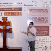 A imagem destaca uma explicação realizada no Museu das Missões diante de uma grande cruz missioneira em madeira exposta sobre base. Uma pessoa fala ao microfone enquanto aponta para elementos do painel ao fundo, que contém textos e gráficos explicativos sobre o território missioneiro e o significado simbólico da cruz.

