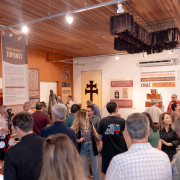 Imagem retrata um espaço interno do Museu das Missões tomado por visitantes, que observam atentamente painéis, esculturas e textos expostos nas paredes. A iluminação destaca os elementos históricos e educativos, criando um ambiente intenso de circulação, troca e interesse pelo patrimônio missioneiro durante a programação do Conecta Missões.

