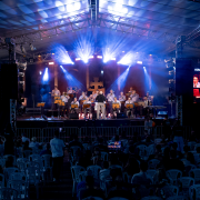 A imagem registra uma visão ampla do palco e da plateia durante a apresentação da Orquestra de Teutônia. O grande telhado metálico da estrutura cobre músicos e público, enquanto luzes de palco iluminam os instrumentistas e dois painéis laterais exibem imagens da apresentação, criando um clima de concerto ao ar livre no coração do Centro Histórico.

