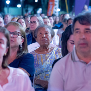 A fotografia apresenta o público sentado em cadeiras organizadas diante do palco, acompanhando atentamente a apresentação musical. Pessoas de diferentes idades aparecem em primeiro plano e ao fundo, sob iluminação artificial noturna, compondo um cenário de participação coletiva e contemplação cultural durante a programação do Conecta Missões.

