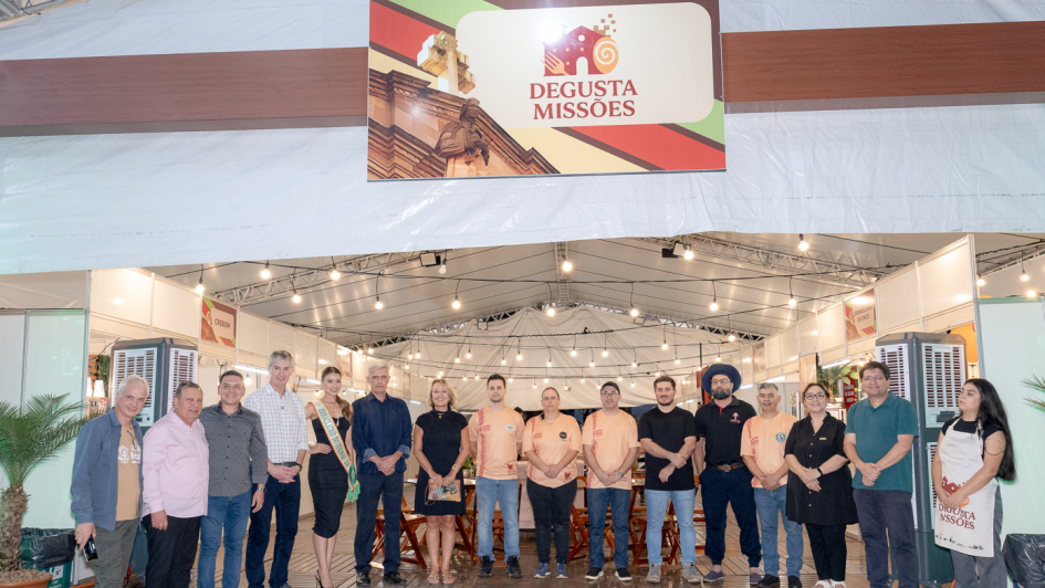 A imagem apresenta um grupo de pessoas posando em frente &agrave; entrada sinalizada do Degusta Miss&otilde;es. Acima, um banner com o nome do evento identifica a aldeia gastron&ocirc;mica, enquanto ao fundo aparecem tendas iluminadas e estruturas do festival, simbolizando a abertura oficial e o car&aacute;ter coletivo da celebra&ccedil;&atilde;o cultural.

