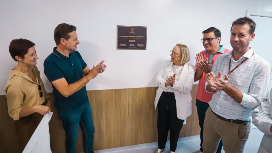 Esta &eacute; uma descri&ccedil;&atilde;o detalhada da imagem para fins de acessibilidade:

A fotografia registra a cerim&ocirc;nia de inaugura&ccedil;&atilde;o da reforma do bloco cir&uacute;rgico e do pronto-socorro do Hospital Santa Terezinha, em Encantado, destacando o momento de celebra&ccedil;&atilde;o das autoridades ap&oacute;s o descerramento da placa.

Elementos Centrais e A&ccedil;&atilde;o
O Grupo: Cinco pessoas (tr&ecirc;s homens e duas mulheres) est&atilde;o posicionadas em semic&iacute;rculo diante de uma parede. Elas est&atilde;o sorrindo e batendo palmas, demonstrando entusiasmo e satisfa&ccedil;&atilde;o com a entrega da obra.

A Autoridade em Destaque: No centro do grupo, uma mulher de cabelos loiros curtos, vestindo um blazer branco elegante sobre uma blusa clara e cal&ccedil;a preta, lidera o aplauso voltada para os demais.

Participantes Laterais: * &Agrave; esquerda, um homem de camiseta polo verde-petr&oacute;leo e cal&ccedil;a jeans, e uma mulher com camisa bege.

&Agrave; direita, dois homens &mdash; um de camiseta polo rosa e outro de camisa social quadriculada clara com crach&aacute; no pesco&ccedil;o.

A Placa de Inaugura&ccedil;&atilde;o: Fixada na parede branca, acima do painel de madeira, h&aacute; uma placa retangular preta com letras douradas. No topo, destaca-se o bras&atilde;o do Estado do Rio Grande do Sul. O texto menciona a "Inaugura&ccedil;&atilde;o da Reforma do Bloco Cir&uacute;rgico", o programa Avan&ccedil;ar na Sa&uacute;de, o nome do governador Eduardo Leite e da secret&aacute;ria da sa&uacute;de Arita Bergmann.

O Ambiente
Cen&aacute;rio: A sala &eacute; moderna e clean, com a parte superior das paredes em branco e a parte inferior revestida por um painel de madeira clara de tonalidade mel.

Ilumina&ccedil;&atilde;o: A luz &eacute; clara, direta e uniforme, t&iacute;pica de ambientes hospitalares novos, refletindo suavemente nas paredes e no piso.

Composi&ccedil;&atilde;o: A imagem captura o grupo de perfil e meio-perfil, focando na intera&ccedil;&atilde;o humana e no s&iacute;mbolo f&iacute;sico da conquista (a placa).

Atmosfera
A cena transmite um clima de conquista comunit&aacute;ria e avan&ccedil;o na infraestrutura de sa&uacute;de. A linguagem corporal de todos os presentes &mdash; sorrisos abertos e aplausos &mdash; refor&ccedil;a o impacto positivo do investimento estadual para a cidade de Encantado.