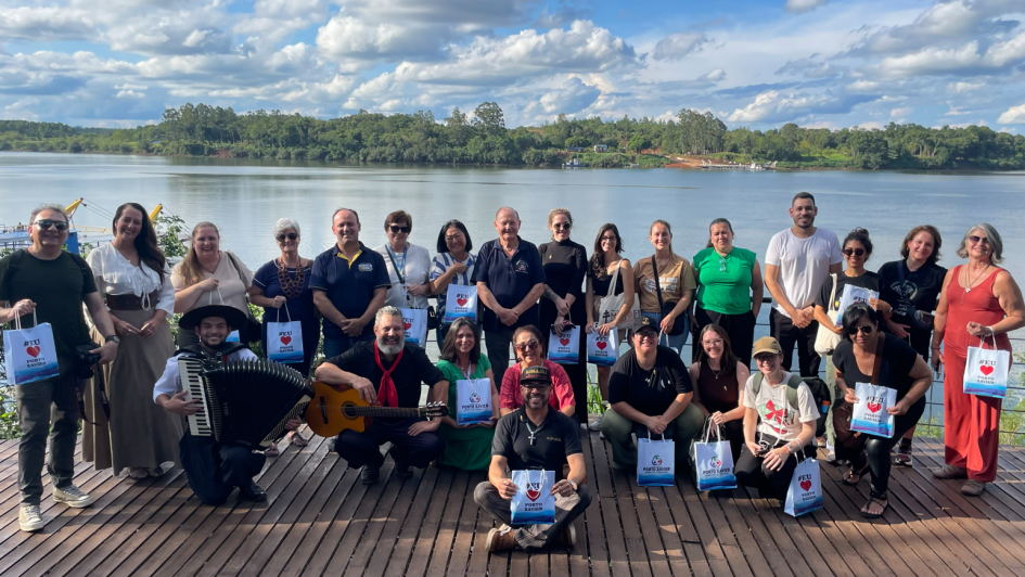 A imagem apresenta um grupo posando sobre um deck de madeira com o Rio Uruguai ao fundo, acompanhados por m&uacute;sicos sentados &agrave; frente com instrumentos. Cada participante segura uma sacola comemorativa, simbolizando o encerramento de uma atividade do Famtour em Porto Xavier, em um ambiente ensolarado e integrado &agrave; paisagem.

