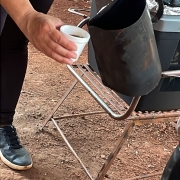 A fotografia registra o preparo e a distribuição do tradicional café de cambona, com uma pessoa servindo a bebida diretamente de uma chaleira de metal escurecida pelo uso, despejando o café em copos pequenos. O cenário é externo, com chão de terra, reforçando o vínculo do ritual com o fogo de chão e a cultura tropeira gaúcha.