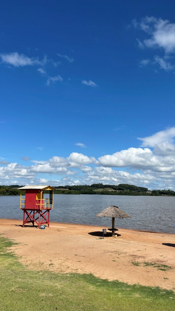 A fotografia apresenta a paisagem da Prainha de Roque Gonzales, com faixa de areia clara, um guarda-sol rústico ao centro e uma torre de salva‑vidas colorida próxima à água. O Rio Uruguai se estende ao fundo, sob um céu azul com nuvens, destacando o cenário natural do balneário.

