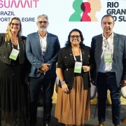 A imagem mostra quatro pessoas alinhadas no palco de um evento, posicionadas diante de um tel&atilde;o com a identidade visual do South Summit Brazil &ndash; Porto Alegre e de um programa institucional do governo estadual. Todas est&atilde;o em p&eacute;, com crach&aacute;s de identifica&ccedil;&atilde;o, e atr&aacute;s delas h&aacute; poltronas claras utilizadas no painel. 