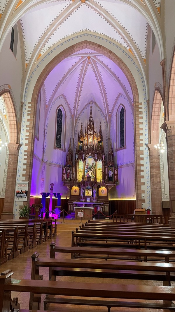 A fotografia mostra o interior da Igreja Matriz Sagrada Família de Nazaré, em Cerro Largo, vista a partir do corredor central entre os bancos de madeira. O altar, ricamente ornamentado e iluminado, destaca imagens sacras e detalhes do estilo gótico, com a nave decorada por arcos altos e luzes suaves.

