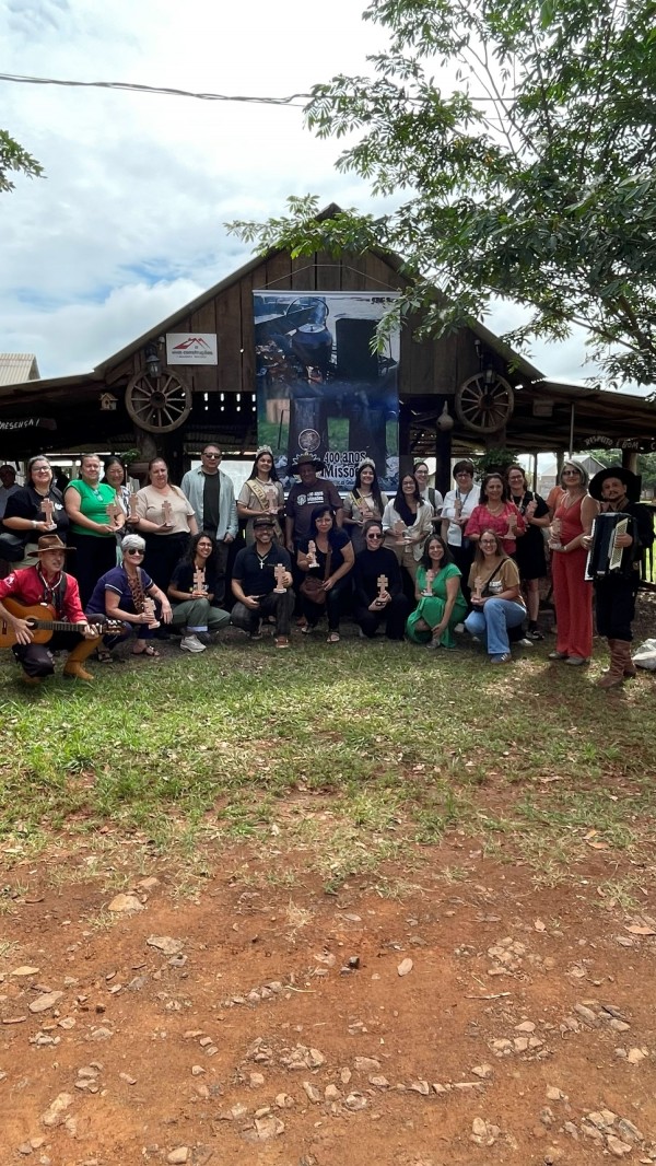 A última imagem retrata o grupo posando em área externa, em frente a uma construção rústica de madeira que remete à arquitetura regional, com um banner alusivo aos 400 Anos das Missões. Músicos aparecem segurando instrumentos, enquanto o terreno de chão batido e a vegetação ao redor reforçam a conexão com a cultura, a história e a identidade missioneira.

