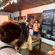A imagem mostra um grupo reunido em uma sala expositiva, onde uma pessoa conduz uma explicação diante de vitrines com objetos arqueológicos e painéis informativos sobre a redução de São Nicolau. Ao fundo, há um grande retrato artístico de um indígena e painéis verticais com textos e imagens históricas, compondo um ambiente fechado, iluminado por trilhos de luz no teto de madeira.