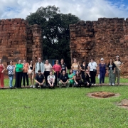 A imagem registra outro momento de grupo posando diante das ruínas missioneiras, com enquadramento mais próximo das pessoas e das paredes de pedra ao fundo. O gramado verde e o céu claro reforçam o caráter aberto e preservado do sítio arqueológico.