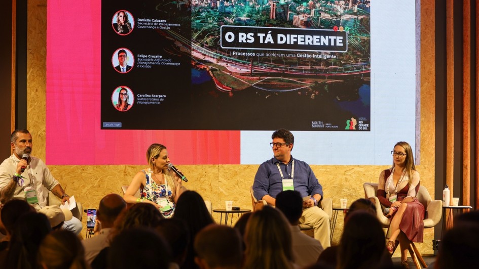 Esta &eacute; uma descri&ccedil;&atilde;o detalhada da imagem para fins de acessibilidade:

A fotografia registra um painel de discuss&atilde;o no palco do RS Innovation Stage, durante o South Summit Brazil 2026, focado em gest&atilde;o p&uacute;blica inteligente.

O Palco e os Participantes
Os Painelistas: Quatro pessoas (dois homens e duas mulheres) est&atilde;o sentadas em poltronas girat&oacute;rias cinzas sobre o palco.

&Agrave; esquerda: Um homem de barba e camisa branca segura um microfone e observa os colegas.

Ao centro-esquerda: Uma mulher loira, vestindo uma blusa estampada colorida, fala ao microfone.

Ao centro-direita: Um homem de &oacute;culos e camisa azul marinho ouve a fala, sorrindo levemente.

&Agrave; direita: Uma mulher de &oacute;culos e vestido bord&ocirc; observa a plateia.

Cenografia: O palco possui uma base de madeira compensada (OSB) e as laterais s&atilde;o decoradas com ripas de madeira verticais. Entre os participantes, h&aacute; pequenas mesas de apoio pretas com copos de &aacute;gua.

Conte&uacute;do do Tel&atilde;o
Ao fundo, um grande painel de LED exibe uma apresenta&ccedil;&atilde;o visualmente rica.

T&iacute;tulo Principal: Em destaque, l&ecirc;-se "O RS T&Aacute; DIFERENTE | Processos que aceleram uma Gest&atilde;o Inteligente".

Imagem de Fundo: Uma foto a&eacute;rea de Porto Alegre ao entardecer, com luzes conectadas por linhas digitais, simbolizando uma cidade inteligente.

Identifica&ccedil;&atilde;o dos Palestrantes: &Agrave; esquerda da tela, quatro c&iacute;rculos exibem as fotos e os nomes dos gestores presentes:

Bruno Silveira (Secret&aacute;rio de Habita&ccedil;&atilde;o e Regulariza&ccedil;&atilde;o Fundi&aacute;ria).

Danielle Calazans (Secret&aacute;ria de Planejamento, Governan&ccedil;a e Gest&atilde;o).

Felipe Cruzeiro (Secret&aacute;rio Adjunto de Planejamento, Governan&ccedil;a e Gest&atilde;o).

Carolina Scarparo (Subsecret&aacute;ria de Planejamento).

Logotipos: No canto inferior direito da tela, aparecem as marcas do South Summit e do Governo do Estado do Rio Grande do Sul.

Composi&ccedil;&atilde;o e Atmosfera
Primeiro Plano: A parte inferior da imagem mostra as silhuetas das cabe&ccedil;as de v&aacute;rias pessoas da plateia, indicando que o local est&aacute; cheio.

Ilumina&ccedil;&atilde;o: O ambiente &eacute; bem iluminado, com focos de luz branca no palco e uma luz ambiente quente que destaca o contraste com o brilho do tel&atilde;o.

Clima: A cena transmite inova&ccedil;&atilde;o, transpar&ecirc;ncia e moderniza&ccedil;&atilde;o administrativa, mostrando o di&aacute;logo entre o governo e o ecossistema de tecnologia.