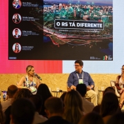 Esta &eacute; uma descri&ccedil;&atilde;o detalhada da imagem para fins de acessibilidade:

A fotografia registra um painel de discuss&atilde;o no palco do RS Innovation Stage, durante o South Summit Brazil 2026, focado em gest&atilde;o p&uacute;blica inteligente.

O Palco e os Participantes
Os Painelistas: Quatro pessoas (dois homens e duas mulheres) est&atilde;o sentadas em poltronas girat&oacute;rias cinzas sobre o palco.

&Agrave; esquerda: Um homem de barba e camisa branca segura um microfone e observa os colegas.

Ao centro-esquerda: Uma mulher loira, vestindo uma blusa estampada colorida, fala ao microfone.

Ao centro-direita: Um homem de &oacute;culos e camisa azul marinho ouve a fala, sorrindo levemente.

&Agrave; direita: Uma mulher de &oacute;culos e vestido bord&ocirc; observa a plateia.

Cenografia: O palco possui uma base de madeira compensada (OSB) e as laterais s&atilde;o decoradas com ripas de madeira verticais. Entre os participantes, h&aacute; pequenas mesas de apoio pretas com copos de &aacute;gua.

Conte&uacute;do do Tel&atilde;o
Ao fundo, um grande painel de LED exibe uma apresenta&ccedil;&atilde;o visualmente rica.

T&iacute;tulo Principal: Em destaque, l&ecirc;-se "O RS T&Aacute; DIFERENTE | Processos que aceleram uma Gest&atilde;o Inteligente".

Imagem de Fundo: Uma foto a&eacute;rea de Porto Alegre ao entardecer, com luzes conectadas por linhas digitais, simbolizando uma cidade inteligente.

Identifica&ccedil;&atilde;o dos Palestrantes: &Agrave; esquerda da tela, quatro c&iacute;rculos exibem as fotos e os nomes dos gestores presentes:

Bruno Silveira (Secret&aacute;rio de Habita&ccedil;&atilde;o e Regulariza&ccedil;&atilde;o Fundi&aacute;ria).

Danielle Calazans (Secret&aacute;ria de Planejamento, Governan&ccedil;a e Gest&atilde;o).

Felipe Cruzeiro (Secret&aacute;rio Adjunto de Planejamento, Governan&ccedil;a e Gest&atilde;o).

Carolina Scarparo (Subsecret&aacute;ria de Planejamento).

Logotipos: No canto inferior direito da tela, aparecem as marcas do South Summit e do Governo do Estado do Rio Grande do Sul.

Composi&ccedil;&atilde;o e Atmosfera
Primeiro Plano: A parte inferior da imagem mostra as silhuetas das cabe&ccedil;as de v&aacute;rias pessoas da plateia, indicando que o local est&aacute; cheio.

Ilumina&ccedil;&atilde;o: O ambiente &eacute; bem iluminado, com focos de luz branca no palco e uma luz ambiente quente que destaca o contraste com o brilho do tel&atilde;o.

Clima: A cena transmite inova&ccedil;&atilde;o, transpar&ecirc;ncia e moderniza&ccedil;&atilde;o administrativa, mostrando o di&aacute;logo entre o governo e o ecossistema de tecnologia.
