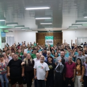 A imagem mostra um grande grupo reunido em um sal&atilde;o amplo, iluminado por luz artificial branca vinda do teto. As pessoas est&atilde;o distribu&iacute;das em p&eacute; por todo o espa&ccedil;o, muitas delas com os bra&ccedil;os erguidos.