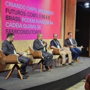 A imagem mostra um painel em um evento, com quatro pessoas sentadas em poltronas alinhadas no palco.