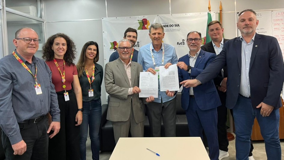 Grupo de nove pessoas lado a lado seguram duas folhas em uma sala. &Agrave; frente, uma mesa vazia com uma caneta. 