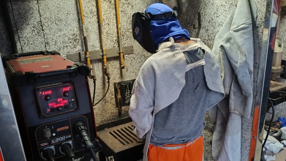 A imagem mostra uma pessoa de costas realizando um trabalho t&eacute;cnico de soldagem em um ambiente industrial ou de oficina. A pessoa est&aacute; equipada com vestimentas de prote&ccedil;&atilde;o e opera um equipamento profissional.

Detalhes do Indiv&iacute;duo e Equipamento
Vestimenta: A pessoa veste uma cal&ccedil;a laranja vibrante e uma camiseta cinza. Por cima, utiliza Equipamentos de Prote&ccedil;&atilde;o Individual (EPIs), incluindo um avental de raspa cinza (couro protetor) que cobre os ombros e as costas, e uma balaclava azul que cobre toda a cabe&ccedil;a e pesco&ccedil;o sob a m&aacute;scara de solda preta, que est&aacute; levantada no momento.

M&aacute;quina de Solda: &Agrave; esquerda, h&aacute; uma m&aacute;quina de solda industrial robusta de cor preta e vermelha, modelo Lincoln Electric C300 CE. O painel digital exibe n&uacute;meros em vermelho (indicando 114 e 20.0) e diversas luzes de controle acesas. Cabos pretos grossos conectam a m&aacute;quina &agrave; &aacute;rea de trabalho.

&Aacute;rea de Trabalho: &Agrave; frente da pessoa, h&aacute; uma bancada de metal com uma grelha preta onde o trabalho de soldagem &eacute; realizado. A parede ao fundo tem uma textura rugosa e clara, com tubula&ccedil;&otilde;es e suportes met&aacute;licos fixados.

Ambiente e Contexto
Cen&aacute;rio: O local parece ser uma unidade m&oacute;vel ou uma oficina especializada, com paredes revestidas de material isolante. &Agrave; direita, pendurados na parede, veem-se outros aventais de prote&ccedil;&atilde;o de couro cinza. O ch&atilde;o &eacute; de metal antiderrapante (estilo "piso de &ocirc;nibus" ou chapa xadrez).

Ilumina&ccedil;&atilde;o: A luz &eacute; funcional e focada na &aacute;rea de opera&ccedil;&atilde;o, destacando o contraste entre o laranja da cal&ccedil;a e os tons industriais de cinza e preto do restante da cena.